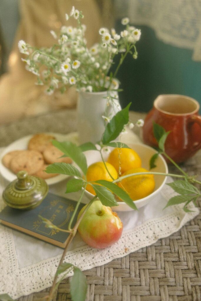 A table featuring a bowl of assorted fruit and a book beside a lemon ricotta cookie.